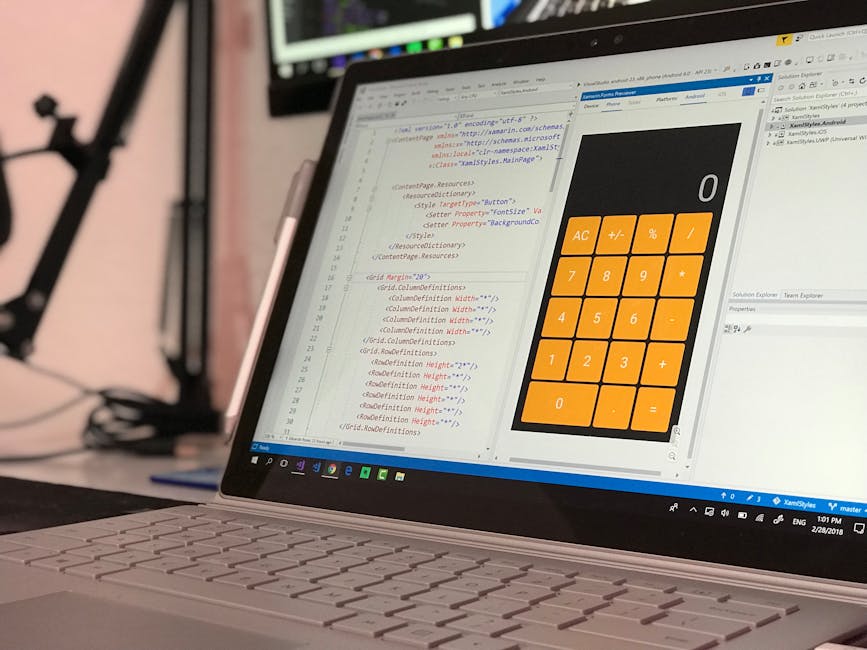 A close-up view of a silver laptop on a desk displaying a software development interface with HTML code on the left side and a large orange calculator application on the right side. The HTML code incl